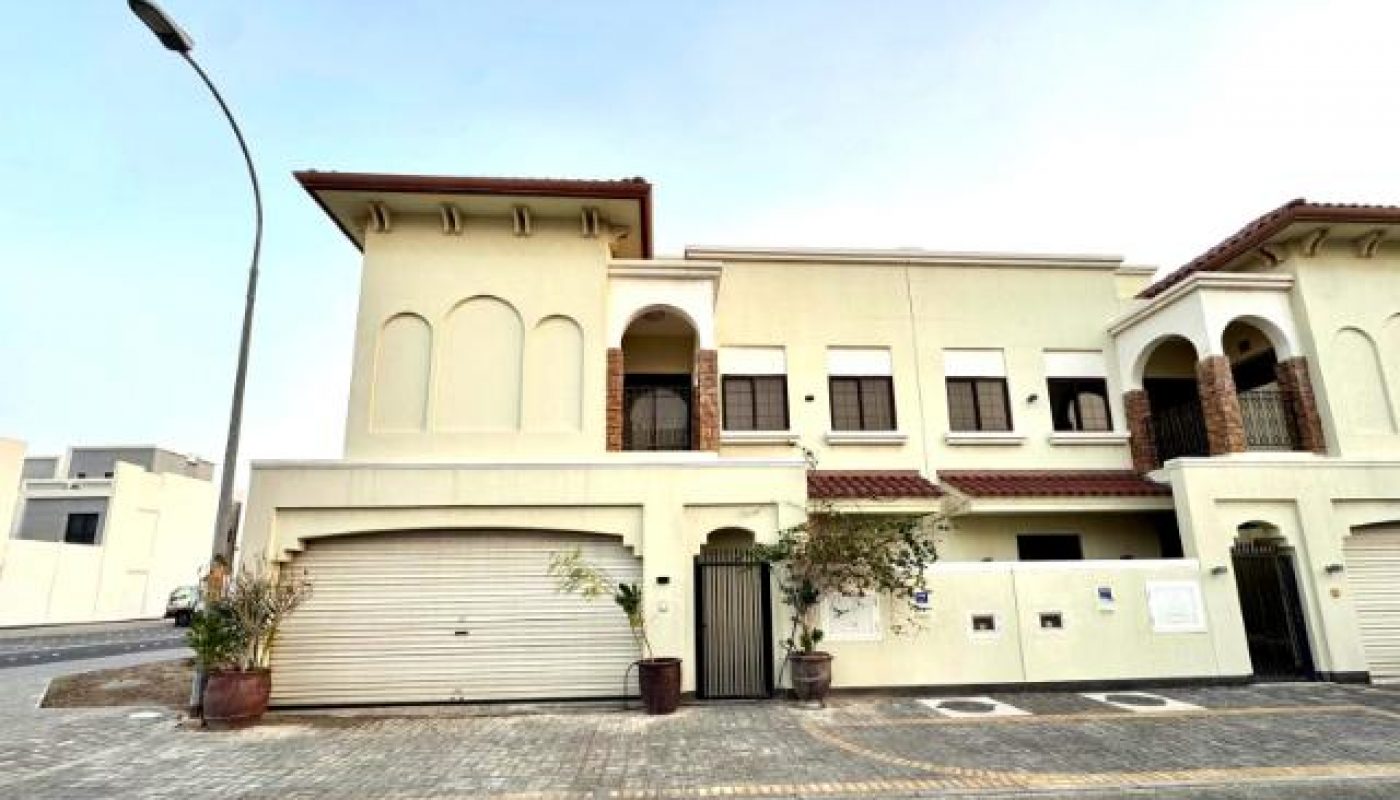 A two-story beige house with arched windows, two garages, potted plants, and a streetlamp, viewed from across a paved street under a clear sky.