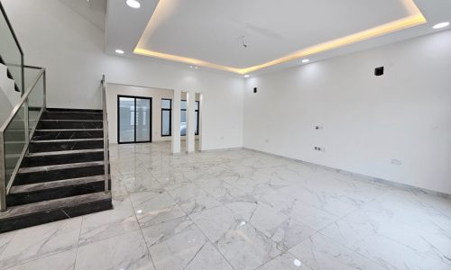 A luxurious, modern villa room featuring marble floors, a staircase with glass railing, recessed ceiling lights, and a large ceiling cut-out with soft lighting. Walls are white with light fixtures and outlets.