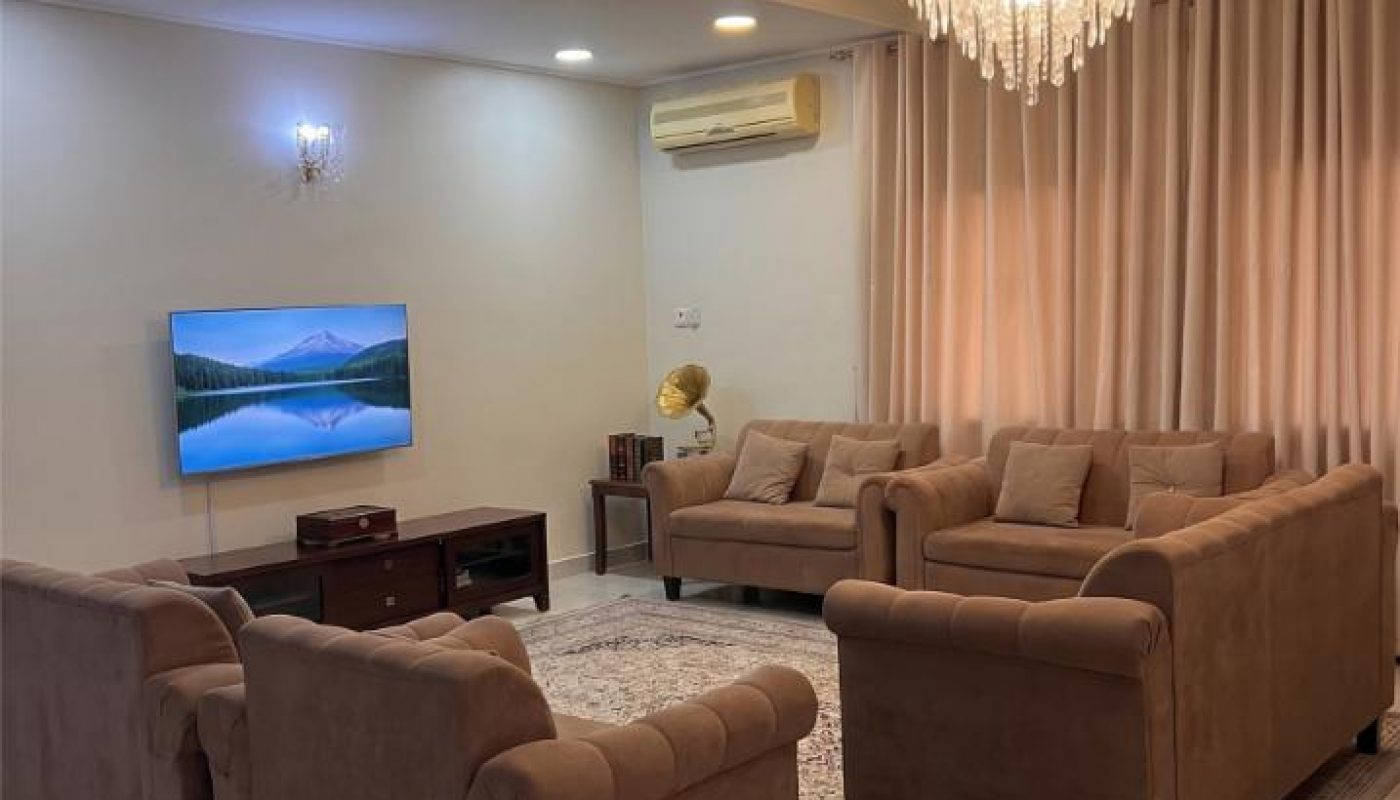 A living room with four brown sofas, a coffee table, TV on a stand, beige curtains, a chandelier, wall sconce, and a split AC unit. The floor is tiled and a rug is under the furniture.