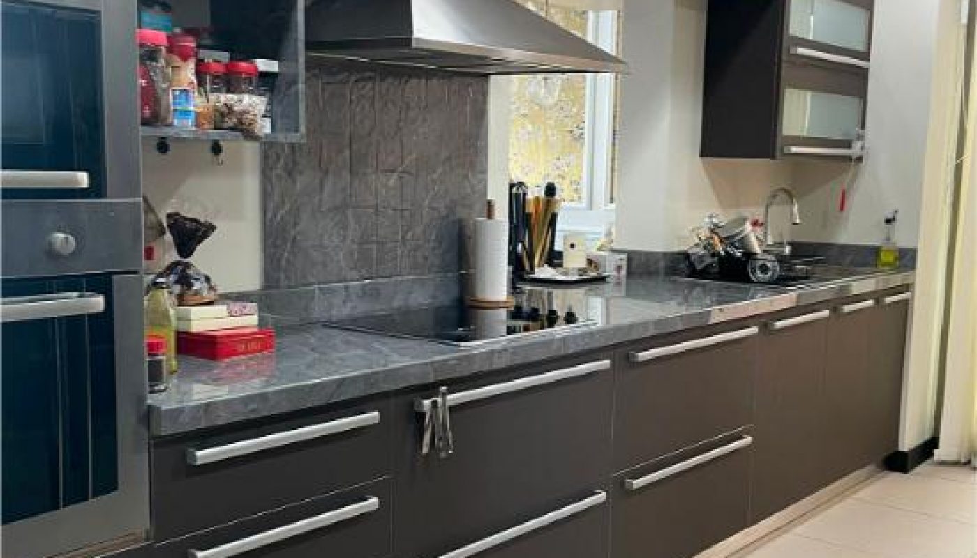 Modern kitchen with dark cabinets, stainless steel appliances, a range hood, countertop items, shelves with jars, and a sink under a window.