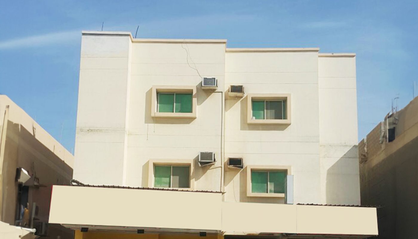 A three-story beige building with two storefronts on the ground floor, green window shades, and multiple air conditioning units on the upper floors.