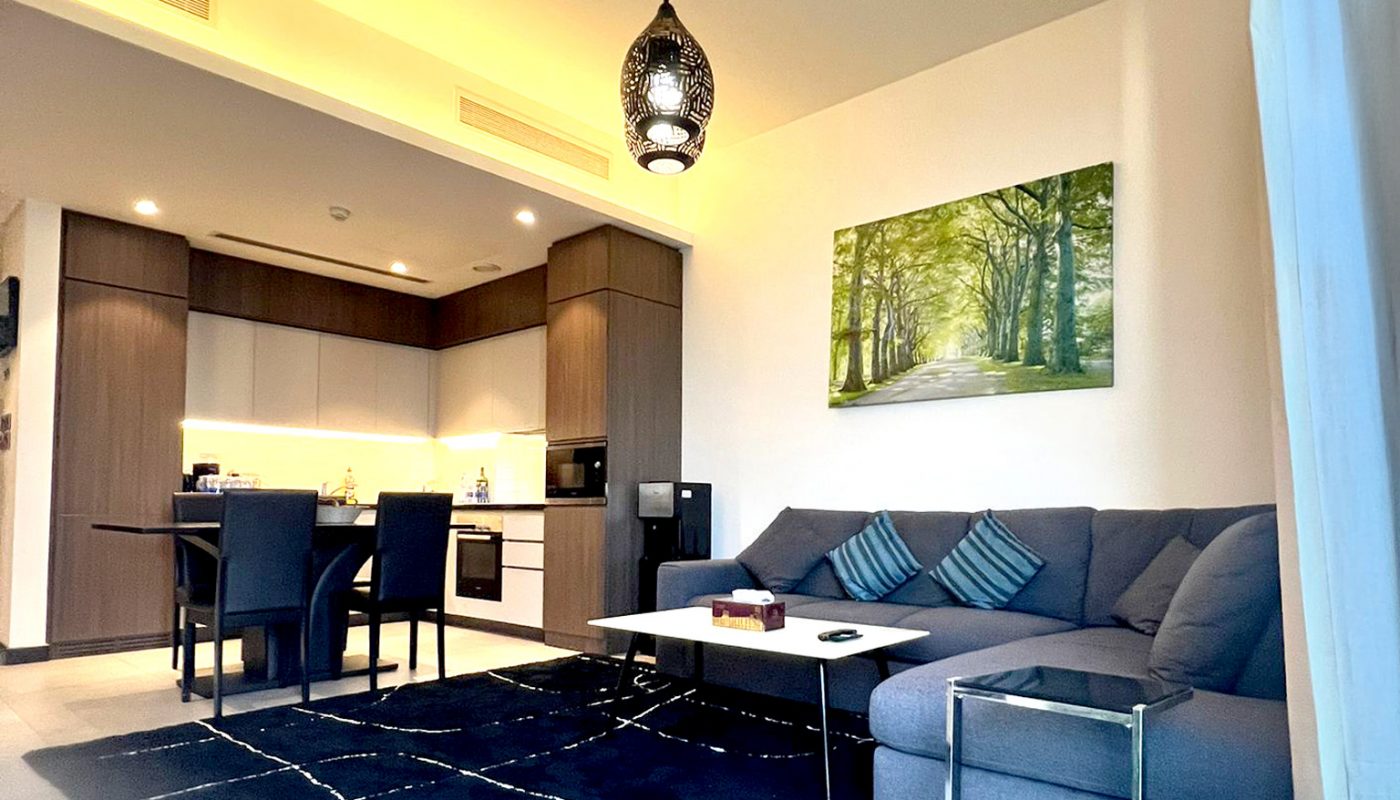 Modern open-plan living room and kitchen with a gray sectional sofa, black area rug, dining area, pendant light, and wall art depicting a forest path.