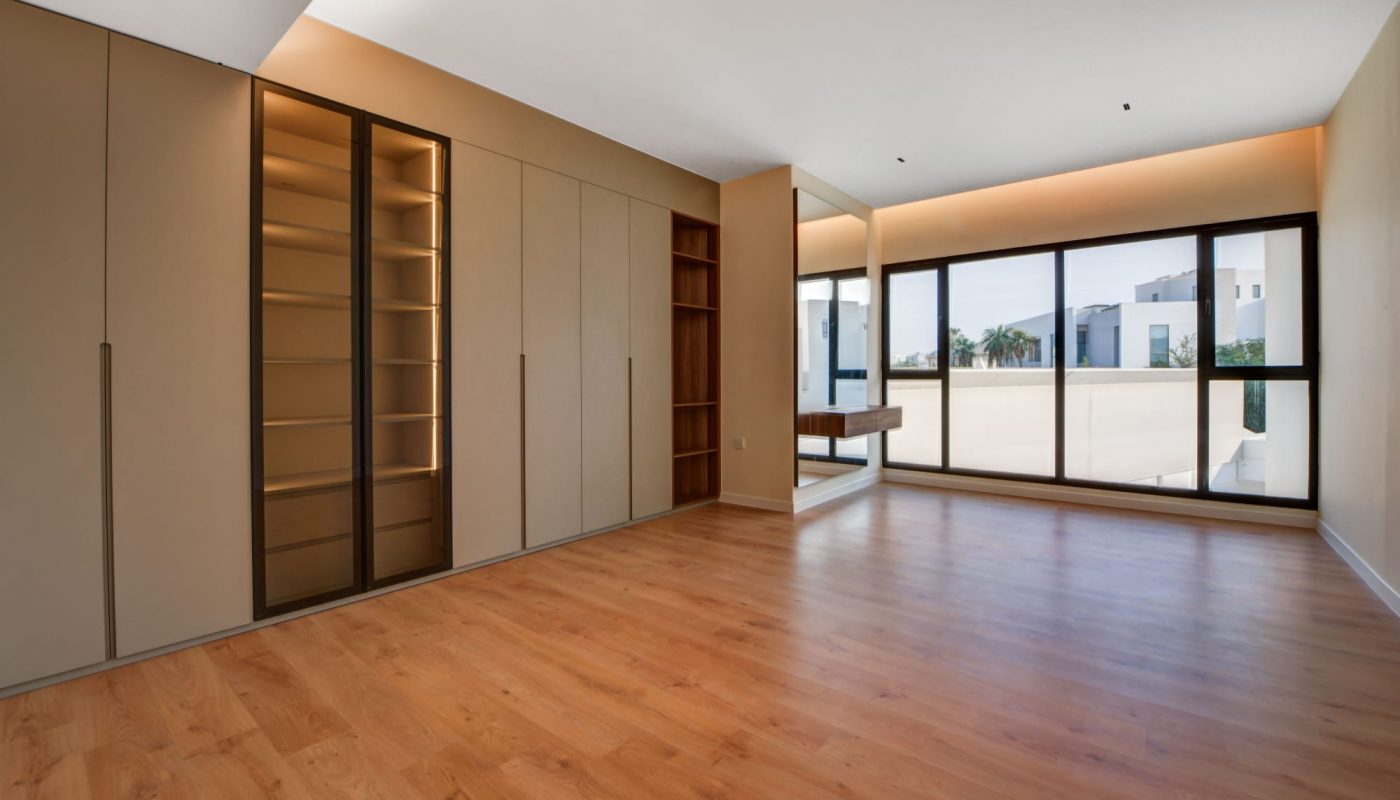 Empty modern room with wooden floor, built-in beige wardrobes, open shelves, large window, and a wall mirror reflecting outdoor buildings and palm trees.