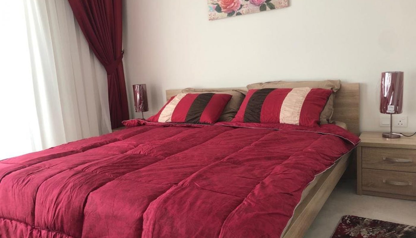 A neatly made bed with red and beige striped pillows and a red comforter, flanked by nightstands and red lamps, with a floral painting above the headboard.