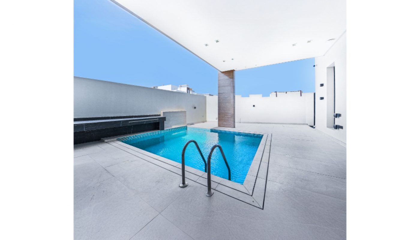 Rectangular outdoor swimming pool with stainless steel ladder, surrounded by white tiles, under a large white canopy, with a bench on the left side and a clear blue sky in the background.
