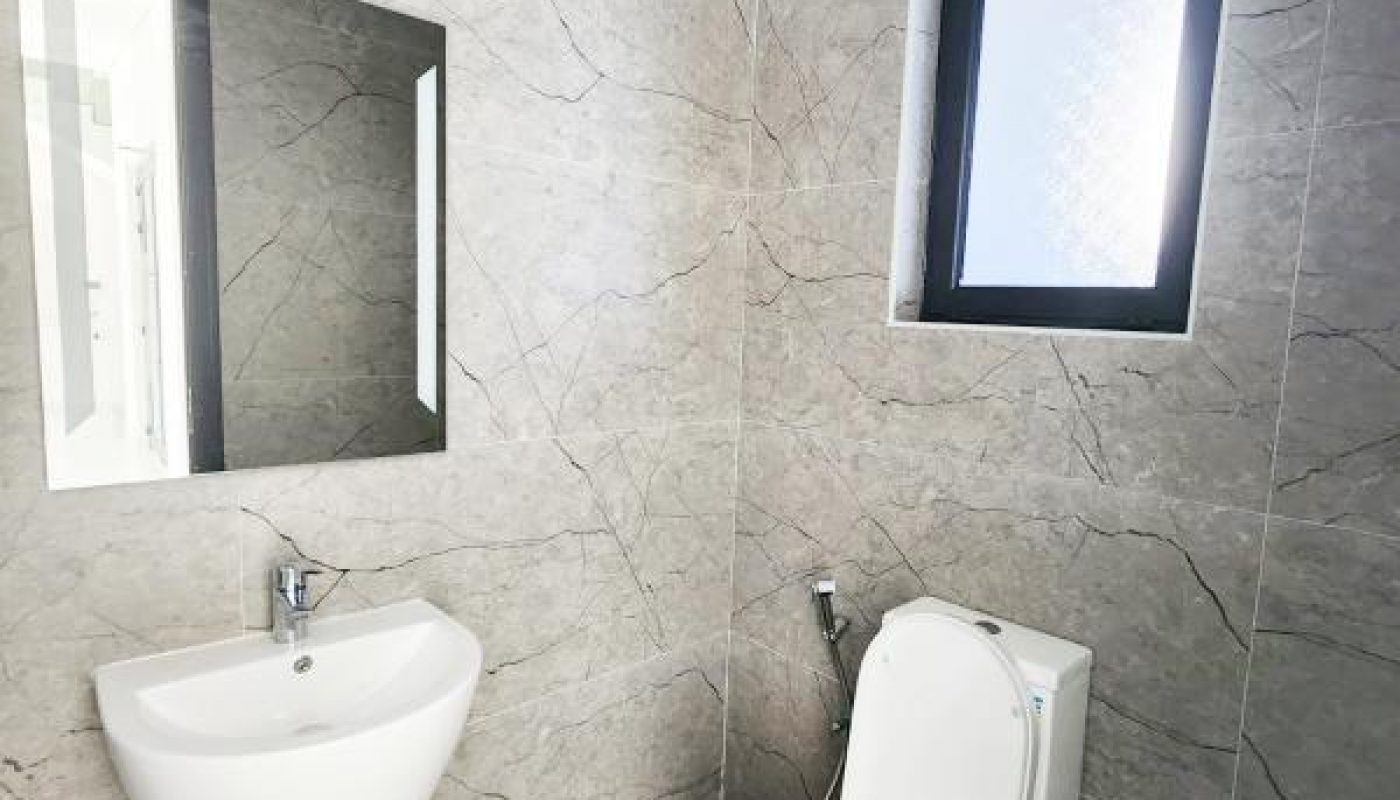 A modern bathroom with a wall-mounted sink, a toilet, a rectangular mirror, and a frosted window. Walls and floor are tiled in gray tones.