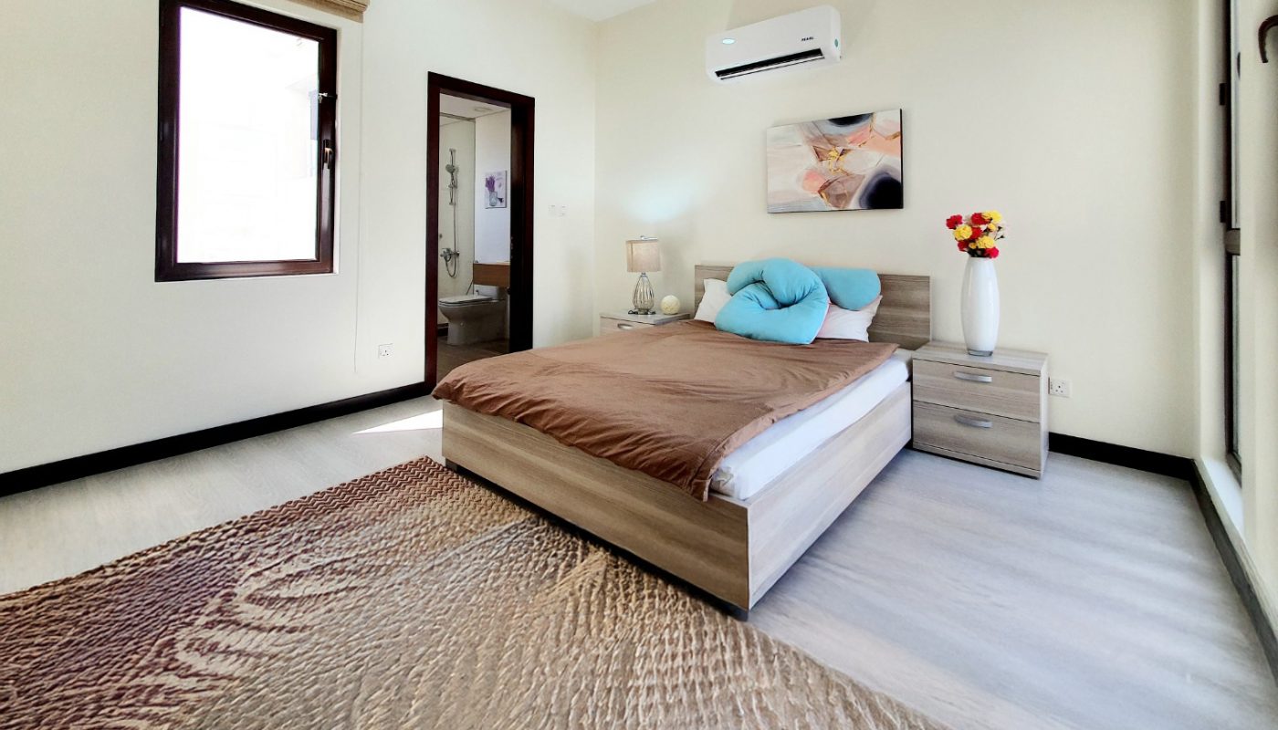 Modern bedroom with a large bed, brown rug, bedside table, vase of flowers, wall art, air conditioner, and large windows letting in natural light.