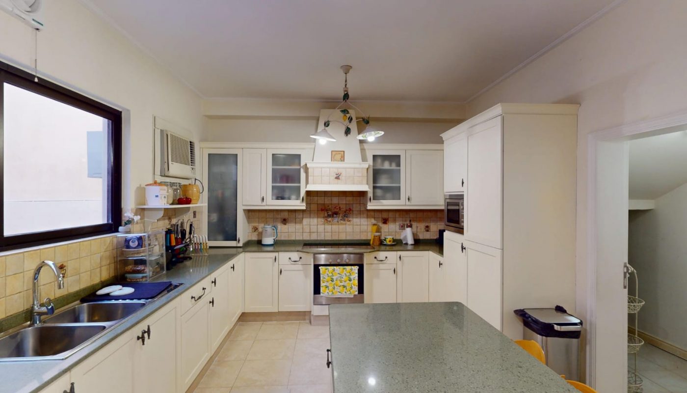 Spacious kitchen with white cabinets, a central island, stainless steel sink, stove, microwave, and various kitchen appliances. Window and storage shelves are on the left wall.