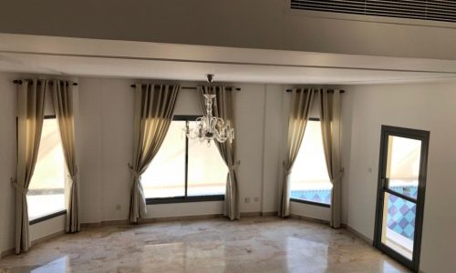 Empty living room with marble floors, draped windows, a chandelier, and a pool view through a door on the right.