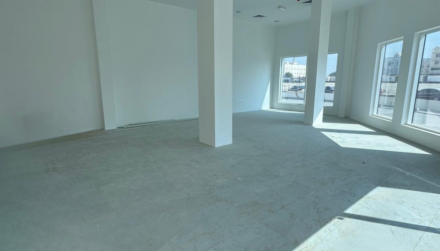 An empty, unfurnished room with white walls, large windows, ceiling lights, and two central support columns. Sunlight streams through the windows onto the floor.