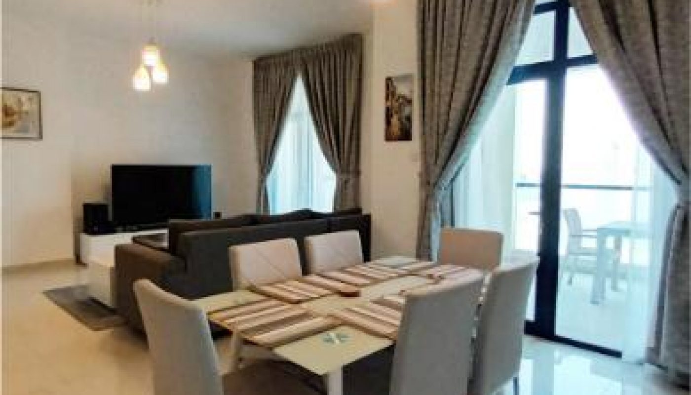 Modern dining and living area with a six-seater table, sofa, TV, large windows, and curtains. Balcony visible through glass door.