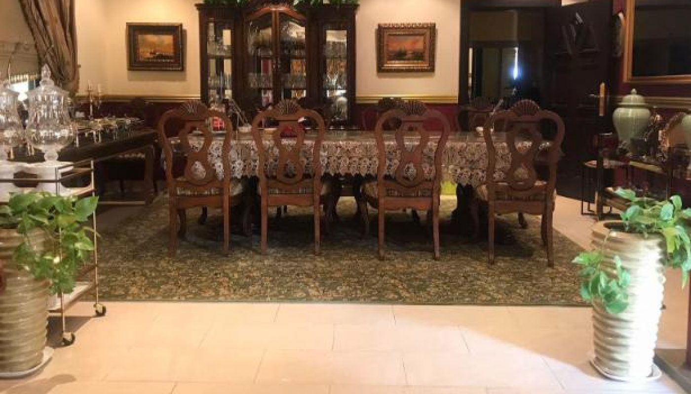 A formal dining room with a long wooden table, ornate chairs, a lace tablecloth, a china cabinet, framed art, and potted plants near the entrance.