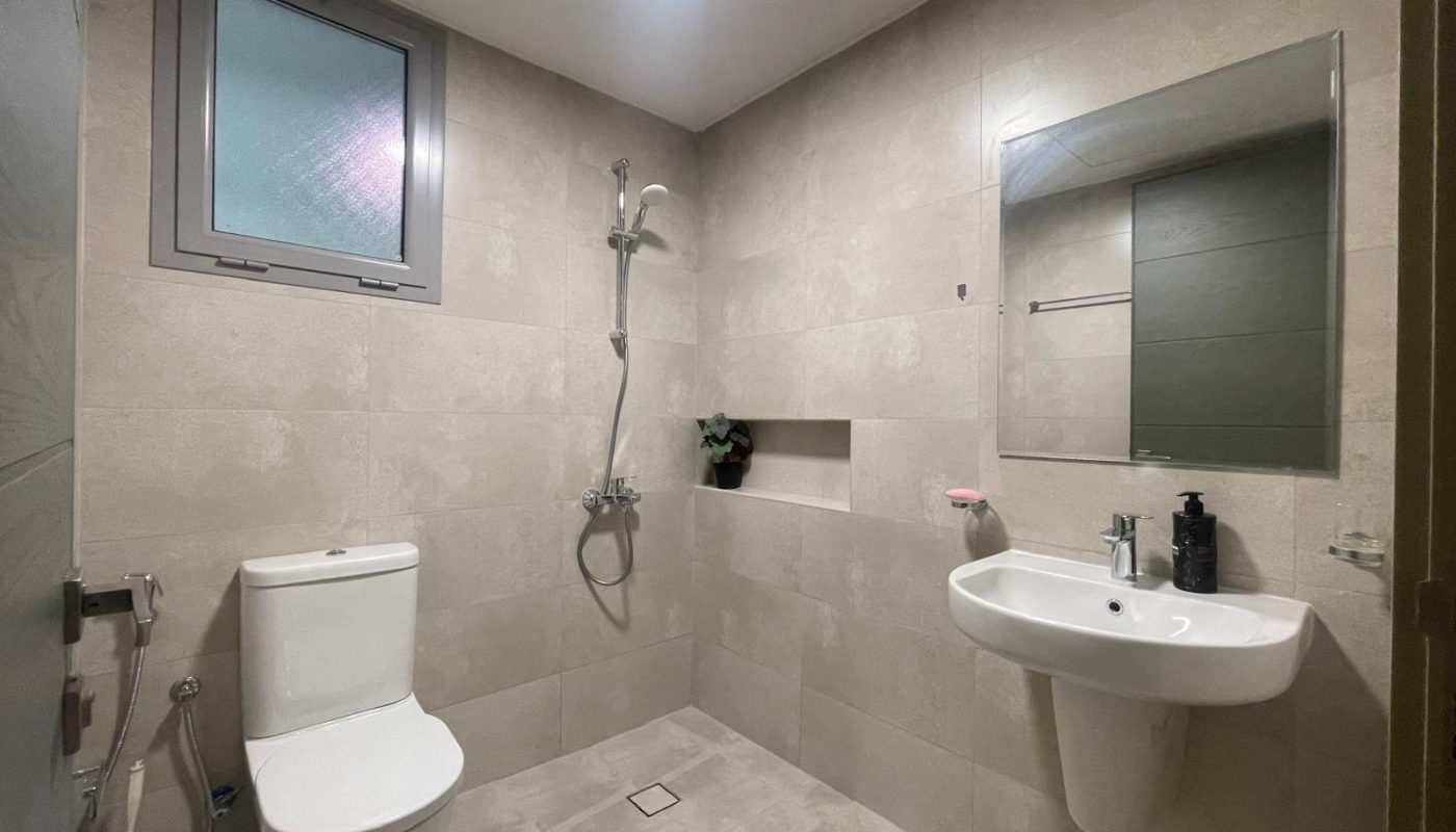 Minimalist bathroom with a toilet, bidet spray, wall-mounted sink, mirror, showerhead, niche shelf, and frosted window. The walls and floor are light beige tiles.
