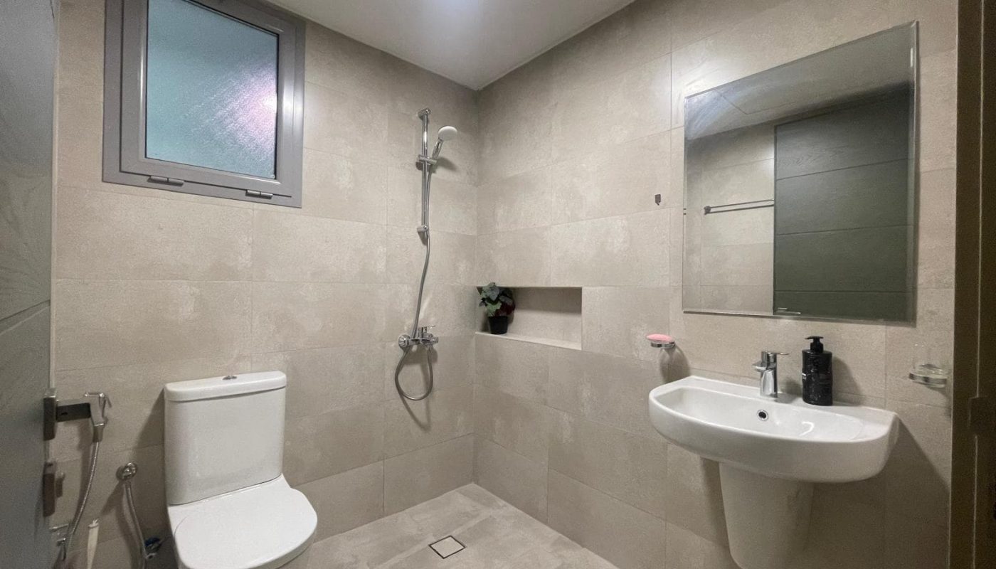 Minimalist bathroom with a toilet, bidet spray, wall-mounted sink, mirror, showerhead, niche shelf, and frosted window. The walls and floor are light beige tiles.