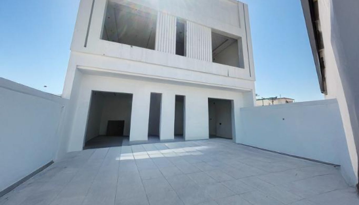 A modern, two-story white building with a large tile courtyard, viewed from a low angle under a bright, sunny sky.
