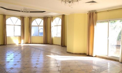 An empty, sunlit room with tiled flooring, multiple arched windows with beige curtains, a sliding glass door, and a ceiling chandelier.