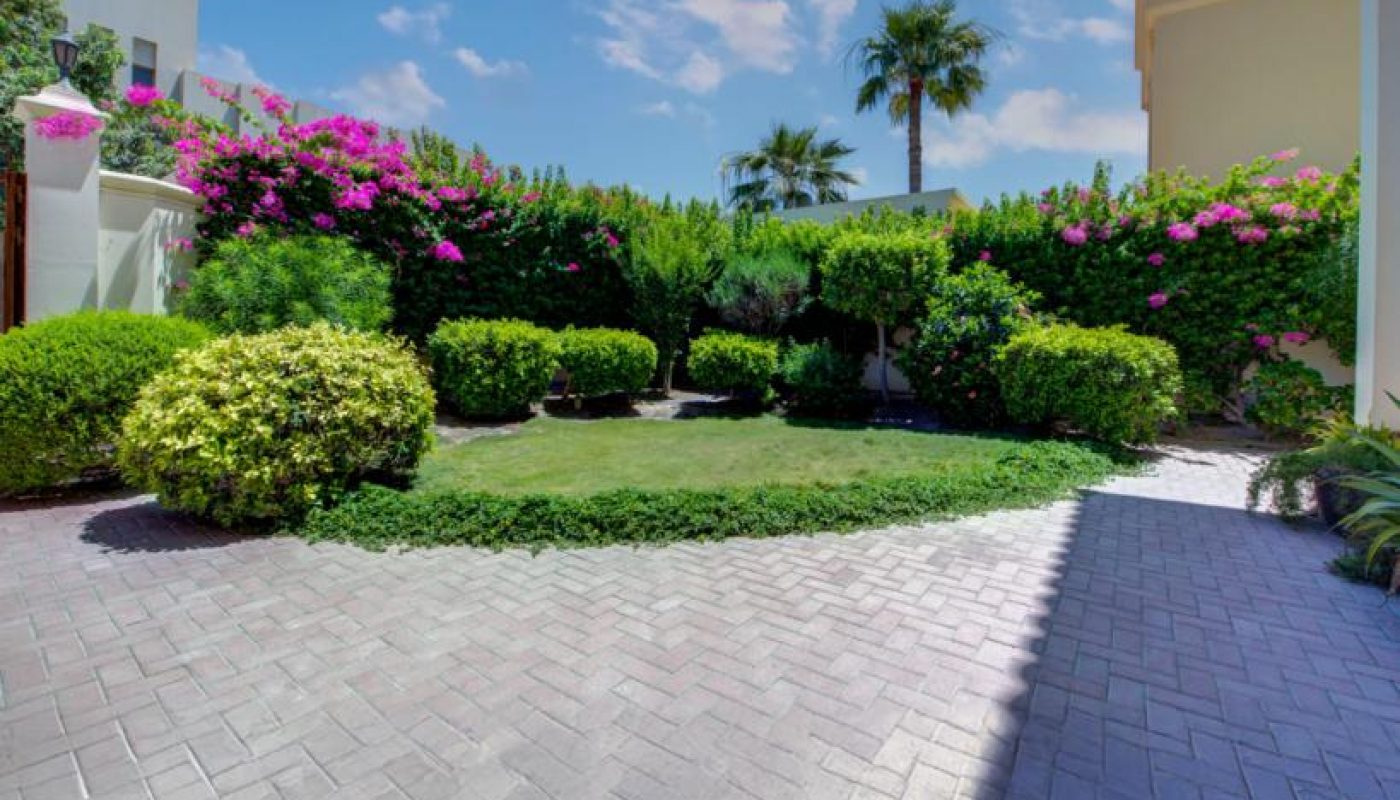 A paved patio area opens to a well-manicured garden with trimmed bushes, flowering plants, and palm trees under a partly cloudy sky.