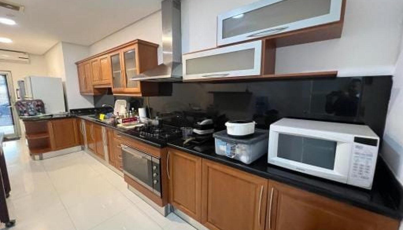 Modern kitchen with wooden cabinets, black countertops, a microwave, oven, and stovetop. White tiled floor. A refrigerator is visible at the end of the room.
