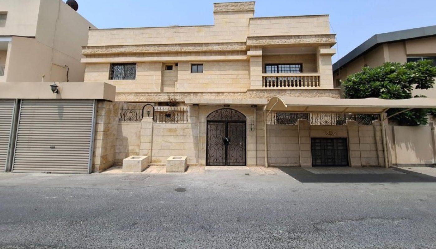 Two-story beige stone house with a central metal gate, small balconies, a garage door on the left, and a carport on the right, set on a quiet street under a clear sky.