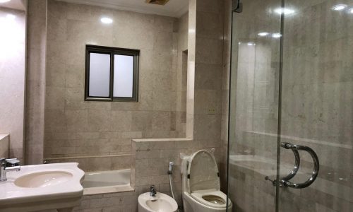 A bathroom with a sink, bathtub, toilet, bidet, and glass-enclosed shower. The walls and floor are tiled in beige. The room has recessed ceiling lights and a small window.