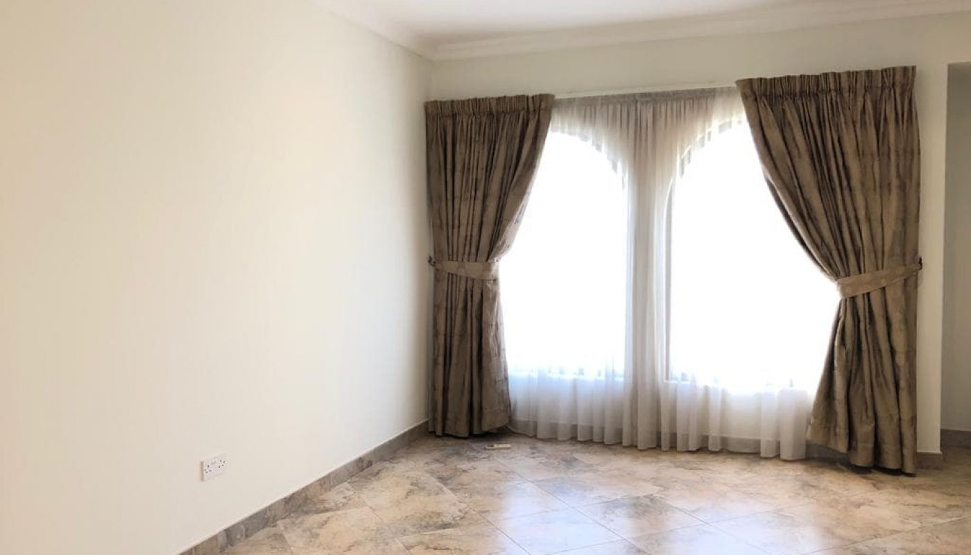A bright, empty room with tiled floor, light-colored walls, and two large windows with double-layered curtains drawn to the sides.