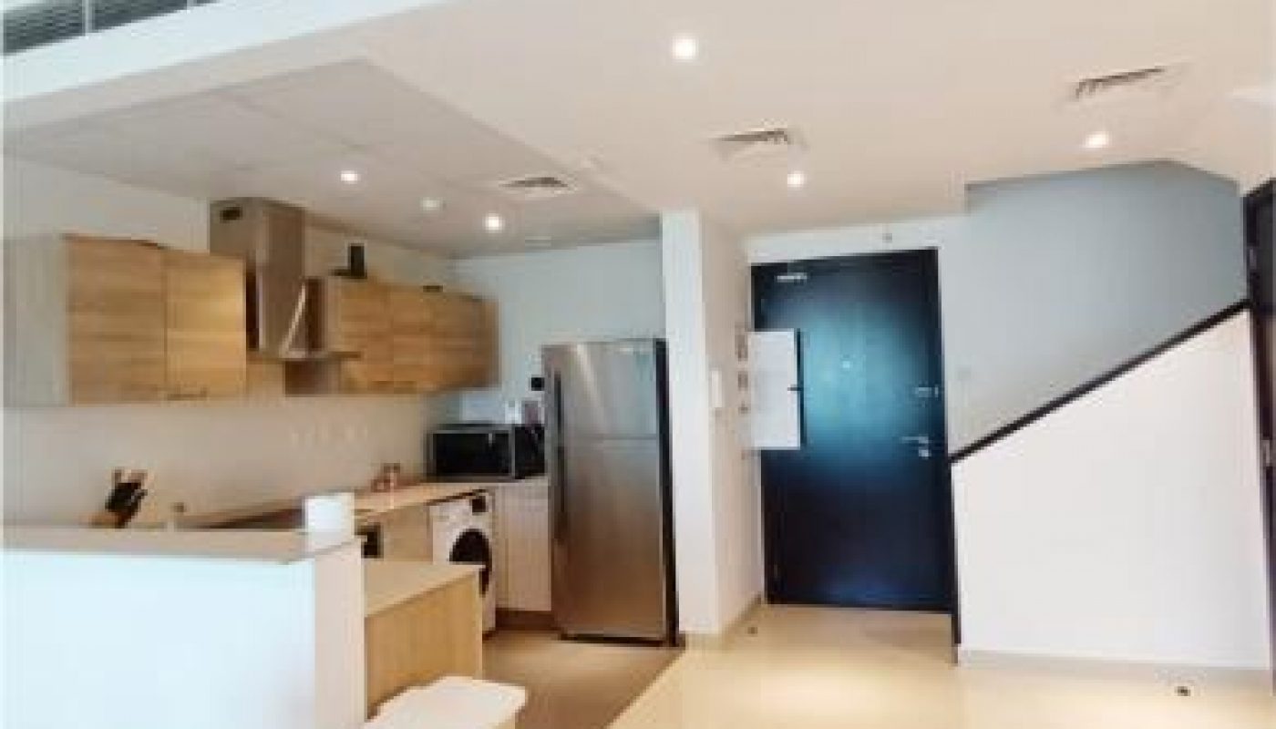 Modern kitchen with light wood cabinets, stainless steel appliances, a trash bin, and white tile flooring, adjacent to a black entrance door and staircase.