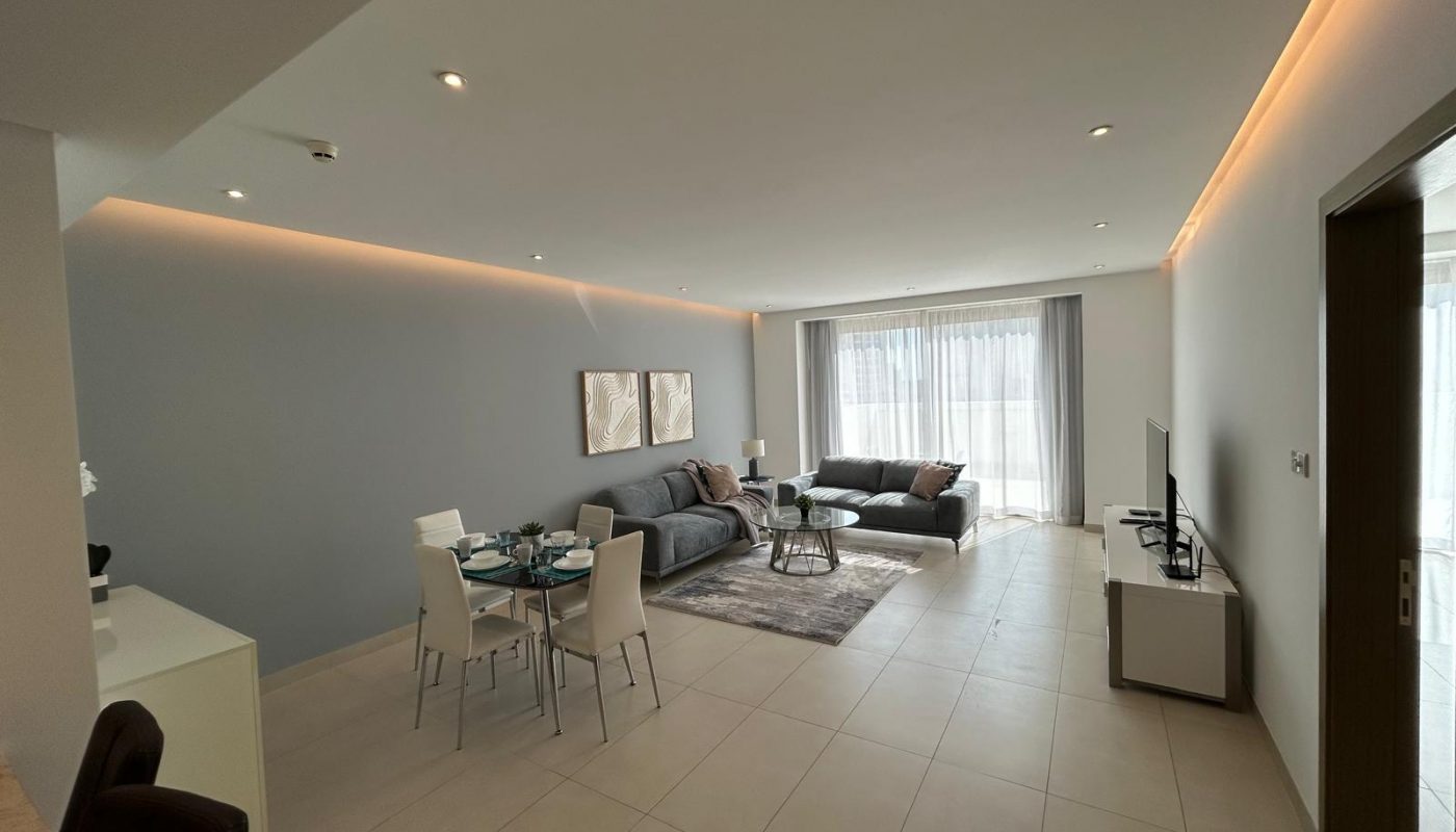 Modern open-plan living and dining area with neutral tones, two sofas, a dining table with four chairs, large windows with sheer curtains, and a wall-mounted TV.