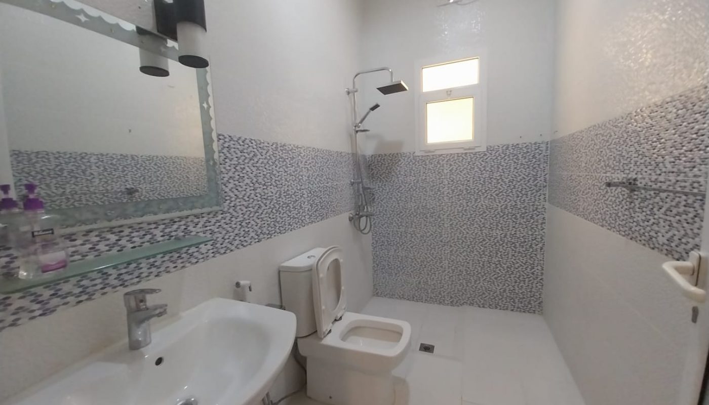 A modern bathroom with a sink, mirror, soap dispensers, toilet, bidet, showerhead, and a small window on the far wall.