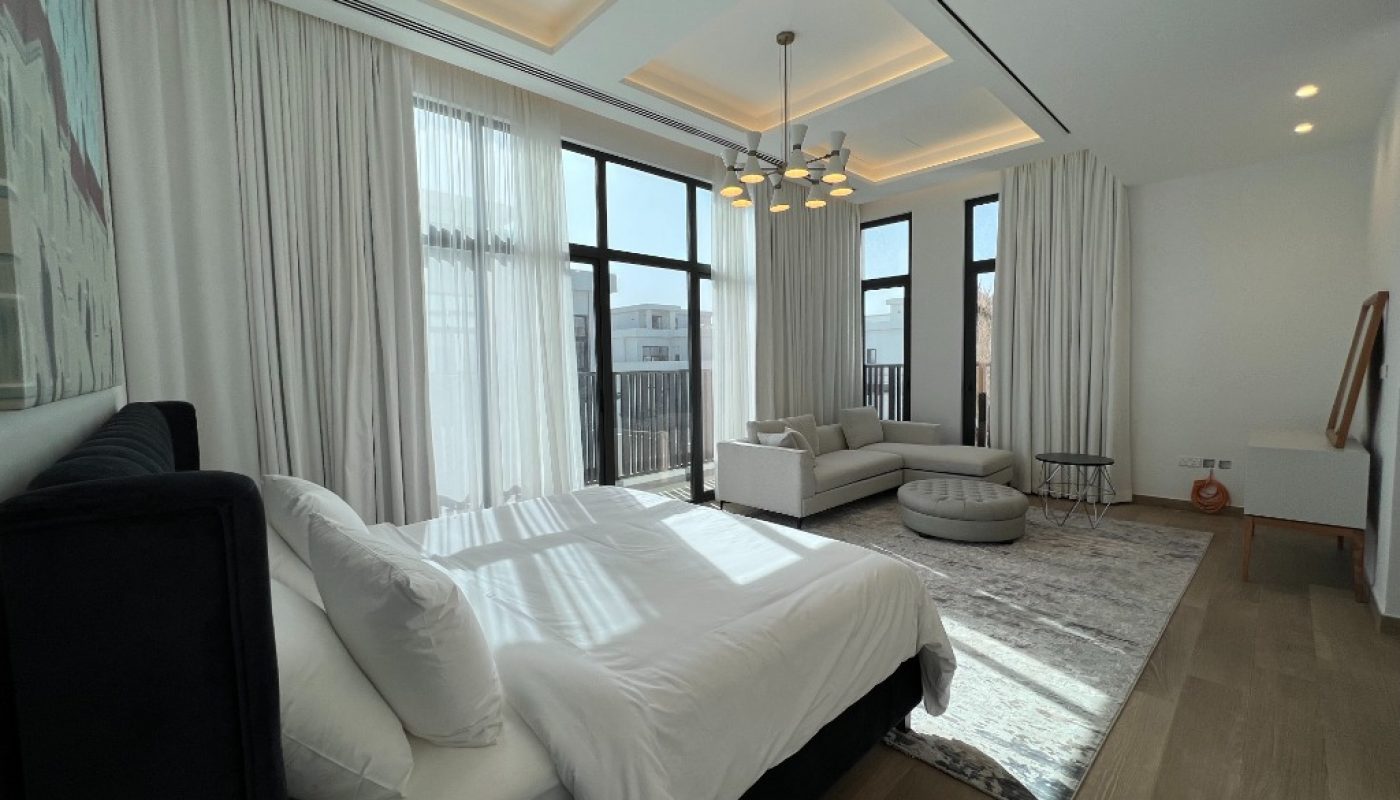 Modern bedroom with a large bed, white bedding, sofa, ottoman, rug, and floor-to-ceiling windows letting in natural light. Light wood flooring and neutral decor complete the space.