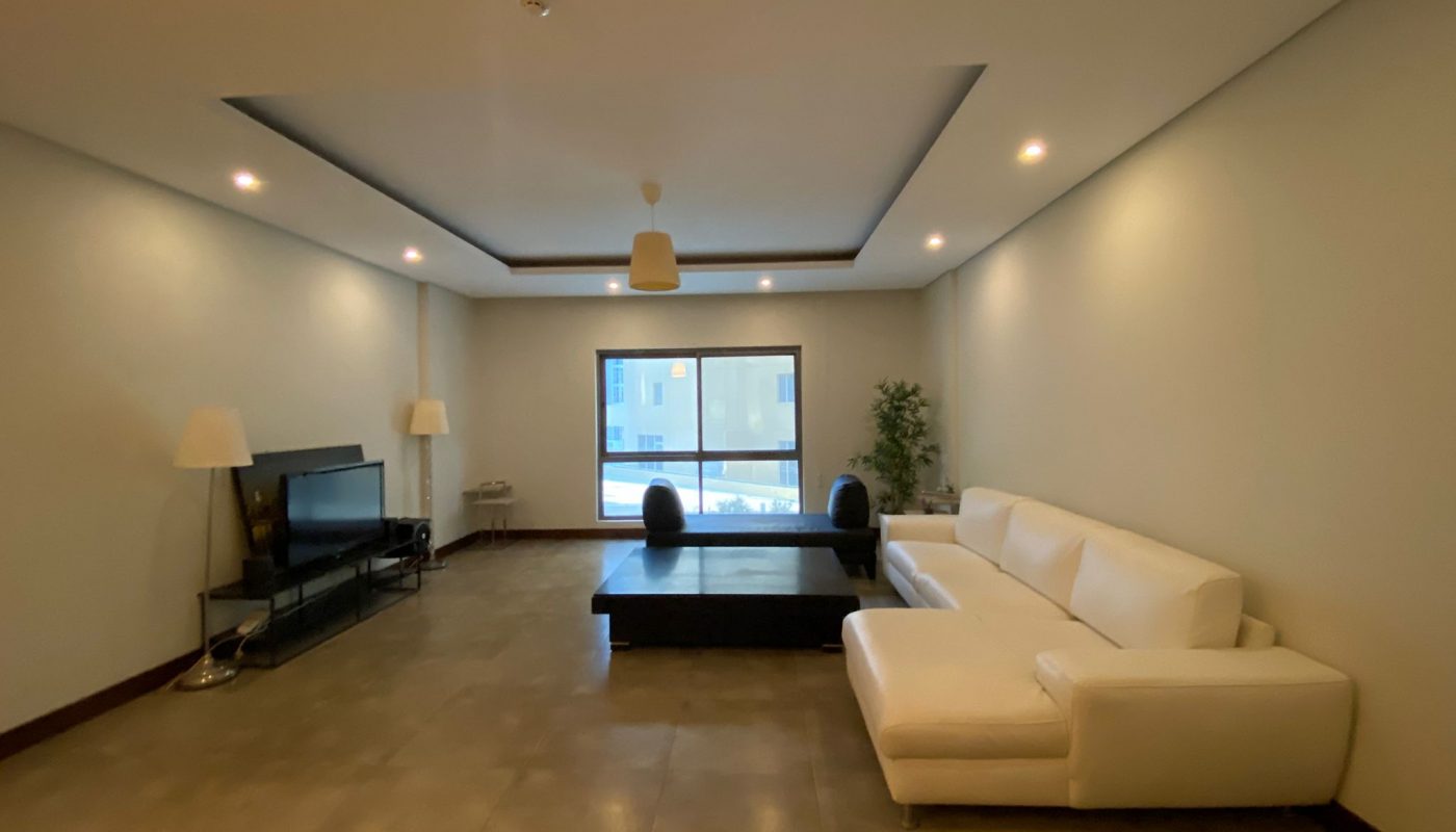 Modern living room with a white sectional sofa, black coffee table, TV on a stand, two lamps, a window, and a potted plant against light-colored walls and a tiled floor.