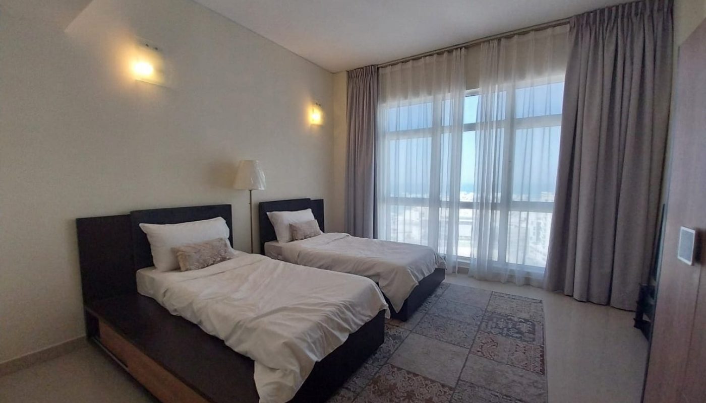 A modern bedroom with two single beds, white bedding, a lamp, large window with sheer curtains, and patterned rug on the floor.
