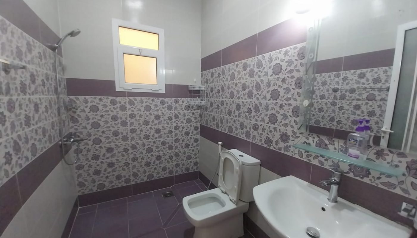 Bathroom with floral-patterned tiles, a toilet, bidet, sink with mirror, wall-mounted soap dispenser, small window, and a shower area without a curtain or partition.
