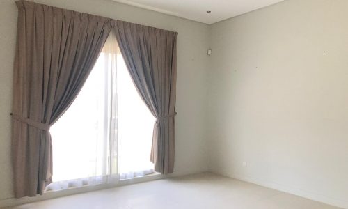 An empty room with a large window covered by brown curtains. The walls are plain and light-colored, the floor is tiled, and there is a ceiling light fixture.