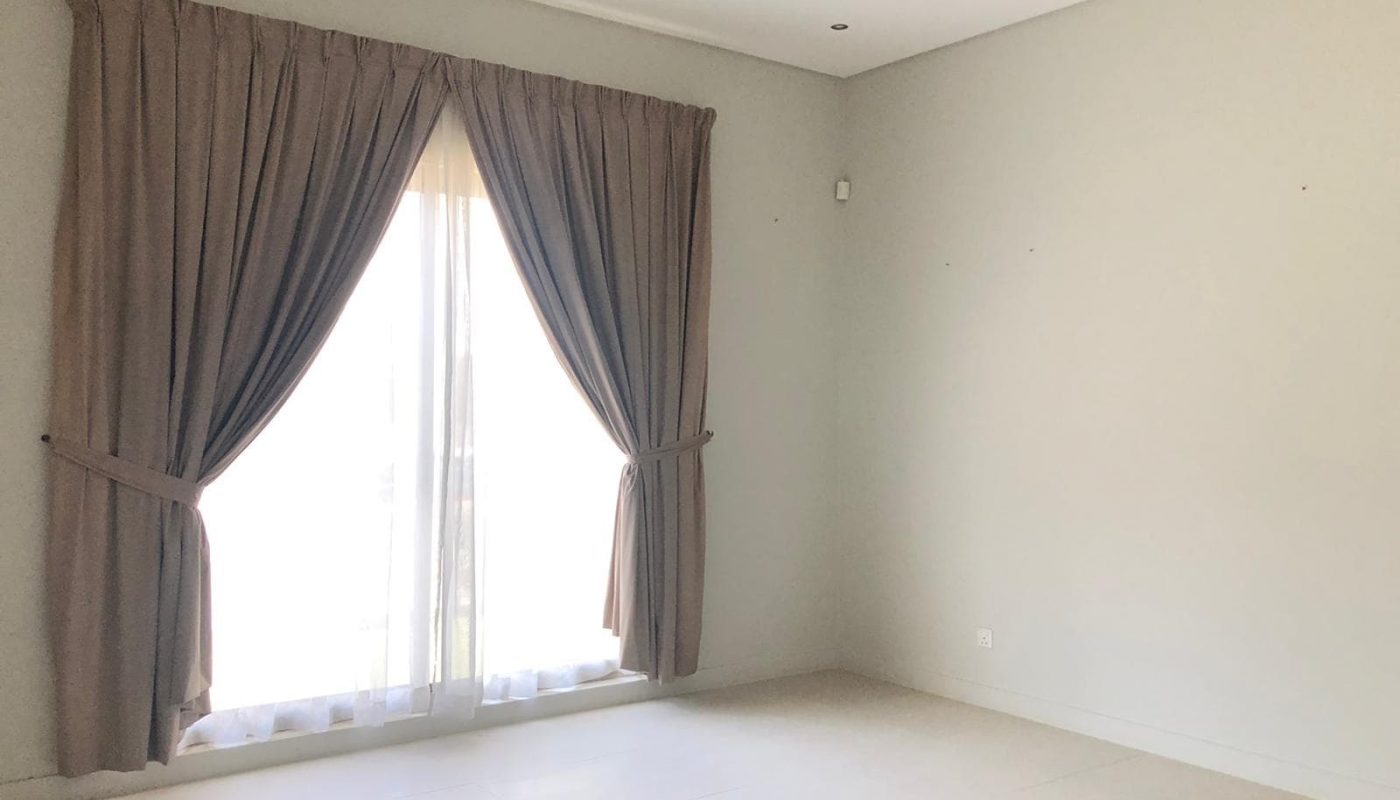 An empty room with a large window covered by brown curtains. The walls are plain and light-colored, the floor is tiled, and there is a ceiling light fixture.