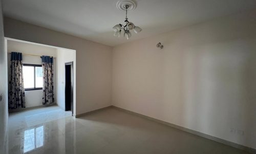 A vacant, unfurnished room with cream-colored walls, a white ceiling light fixture, a window with patterned curtains, and a tiled floor. An adjoining room with a closed door is visible in the background.