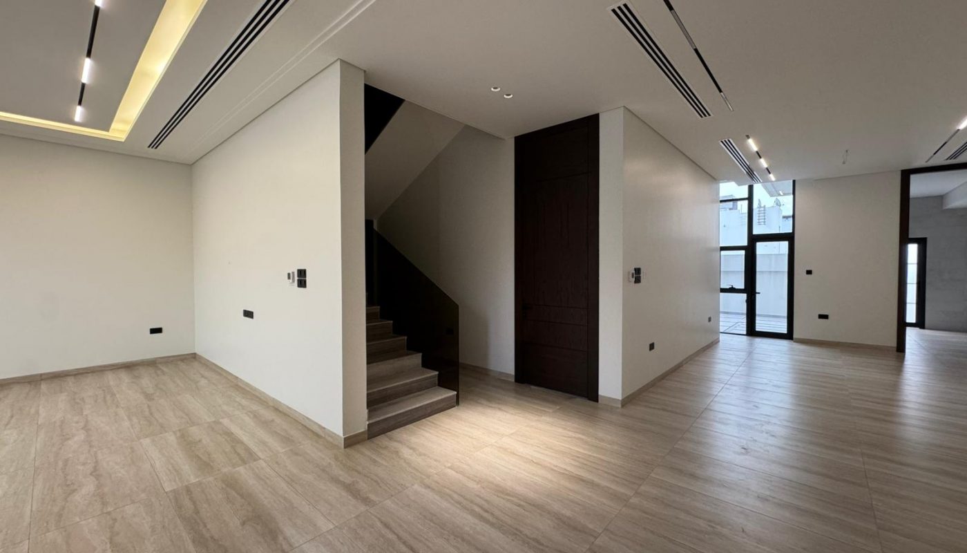 Modern, empty interior space with light wood flooring, white walls, recessed ceiling lights, a staircase, and large windows letting in natural light.