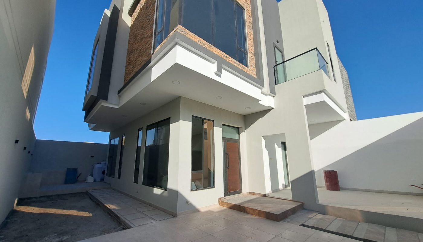 Modern two-story house with large windows, a brick accent wall, flat roof, and tiled front patio under a clear blue sky.