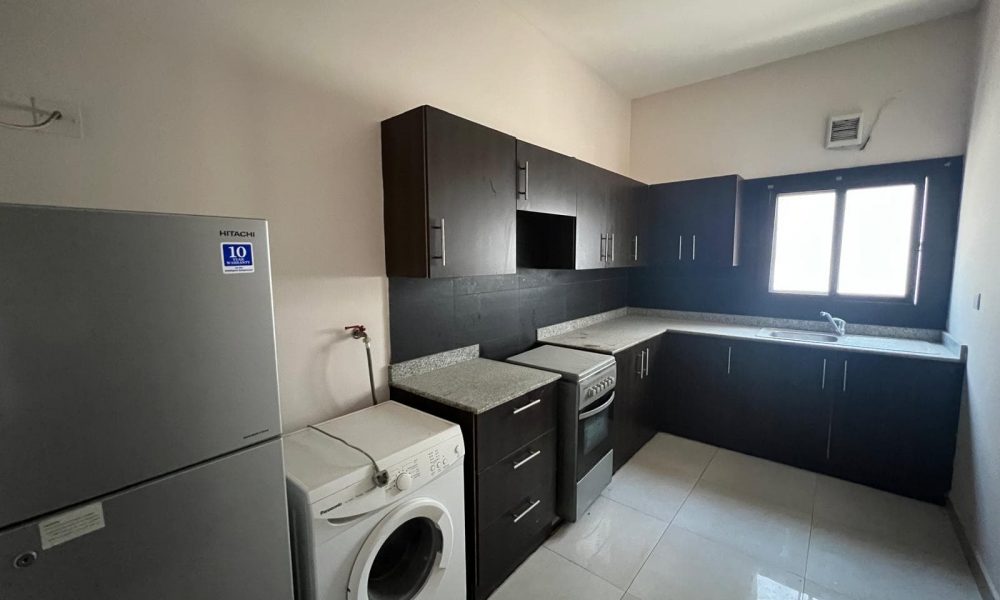 A modern kitchen with dark wood cabinets, a fridge, a washing machine, an oven, and a sink. The room has light-colored walls and a tiled floor. Natural light comes through a window.