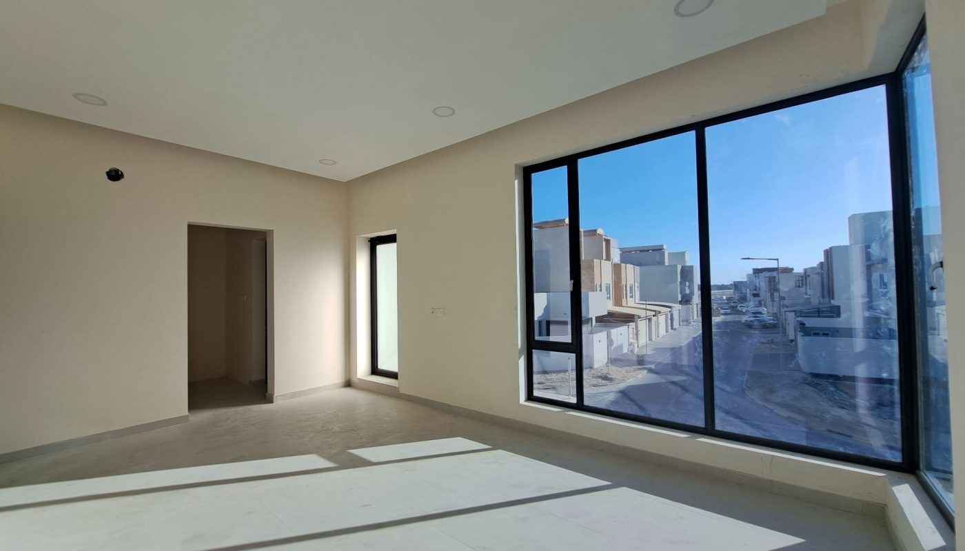 Empty, unfurnished room with large floor-to-ceiling windows showing a sunny street view of modern houses outside. Sunlight casts shadows on the tiled floor.
