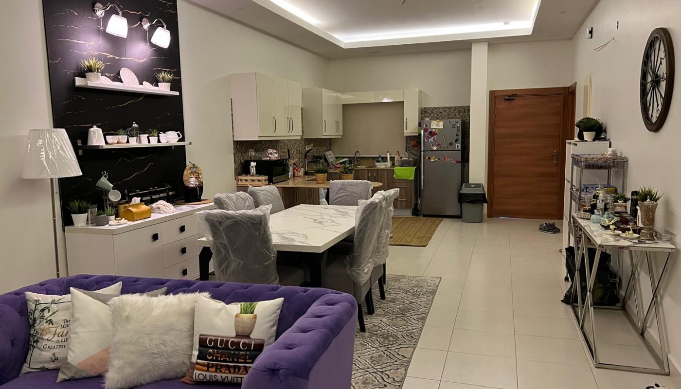 Modern open-plan living and dining area with purple sofa, white dining table, kitchen in the background, and decorative shelves and accessories throughout.