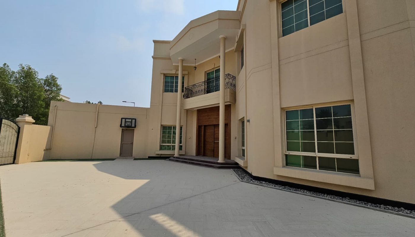 A large, beige two-story house with tall columns, multiple windows, a second-floor balcony, and a spacious paved driveway under a partly cloudy sky.