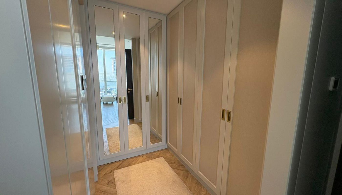 A walk-in closet with mirrored wardrobe doors, built-in cabinets, a beige rug, and a view of a cityscape through a window in the adjacent room.