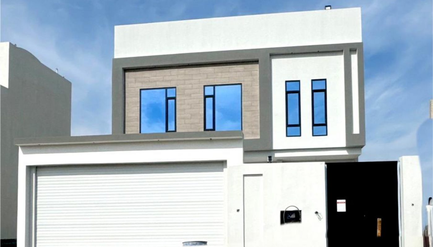 Modern two-story house with a flat roof, large garage door, and rectangular windows, set against a blue sky with scattered clouds.