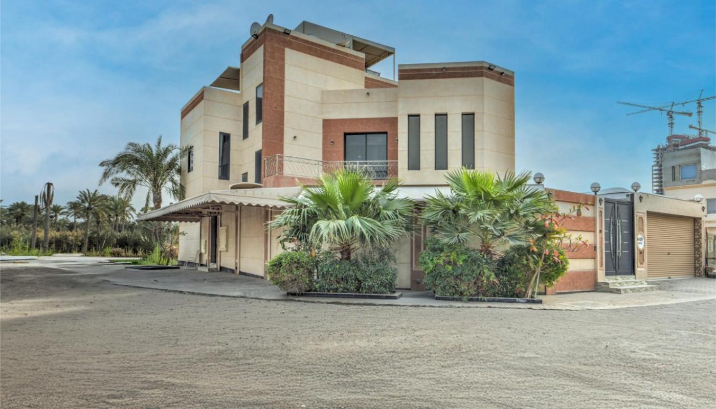 Modern two-story building with beige and reddish-brown exterior, surrounded by palm trees, set against a clear blue sky and paved driveway.