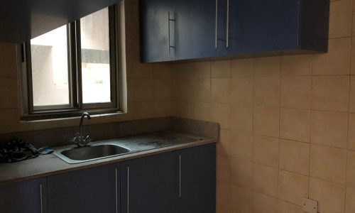 A small kitchen with beige tiled walls, blue cabinets, a stainless steel sink, and a window on the left.