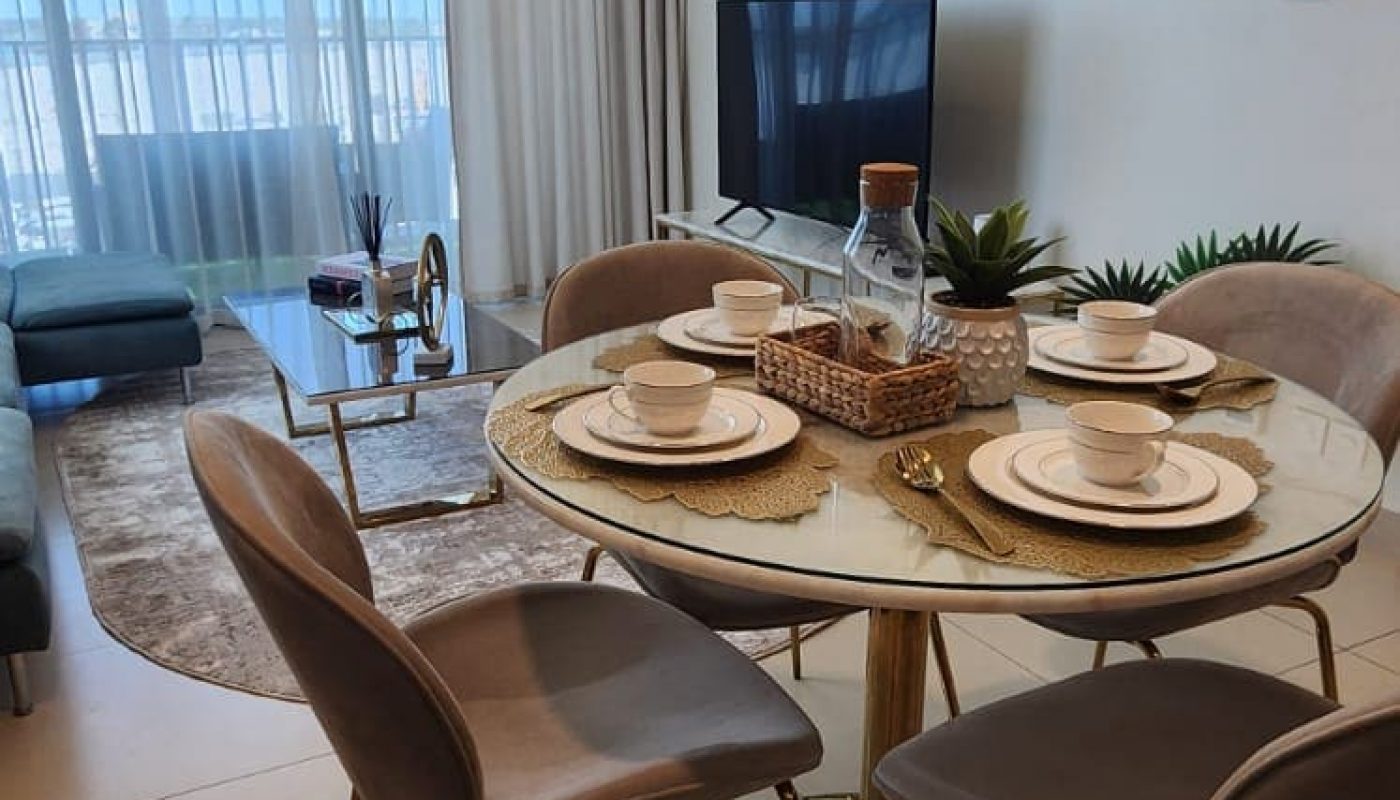 A modern dining area with four chairs around a round table set for four, next to a living area with a TV, couch, coffee table, and large window with sheer curtains.