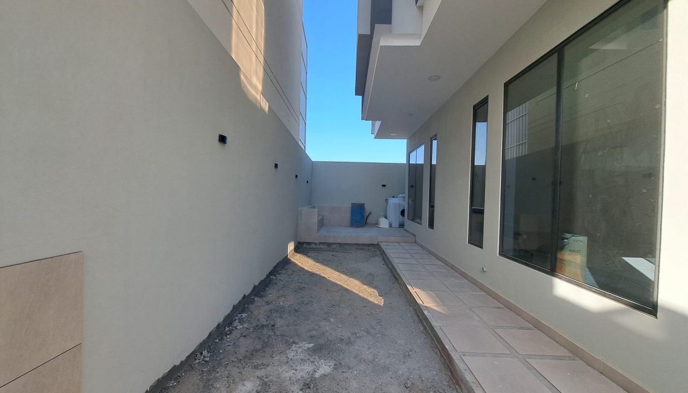 A narrow outdoor walkway beside a modern house under construction, with unfinished ground and tiled path, large windows, and some construction materials at the end.