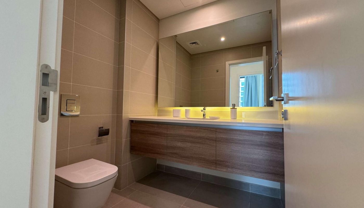 Modern bathroom with beige tiles, wall-mounted toilet, large mirror, wooden vanity, double sinks, and overhead lighting.