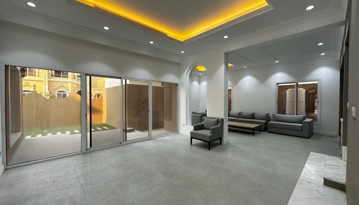 Modern, spacious living room with recessed lighting, large sliding glass doors opening to a small yard, and minimalistic gray furniture against neutral walls and floors.