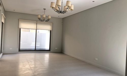 An empty room with light gray walls, tiled flooring, two large windows with blinds, and two chandeliers hanging from the ceiling.